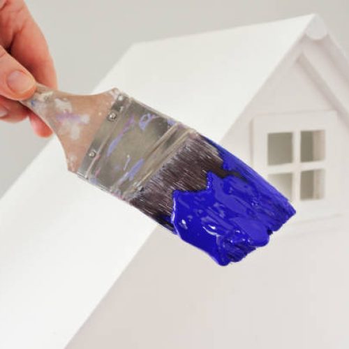 Person holding a paint brush with blue paint color against a white house. Renovation, home decoration and painting house concept. No people. Copy space