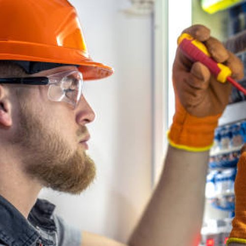 Male electrician working in switchboard. Male electrician in overalls working with electricity. High quality photo. Close-up. Man wearing safety glasses and hard hat.